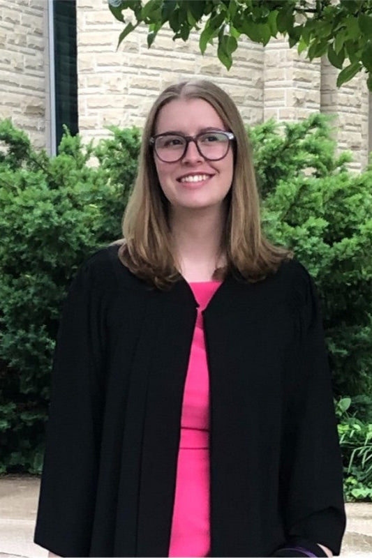 Mary Hamilton wearing a black coat over a pink shirt, standing outdoors with greenery and a building in the background.