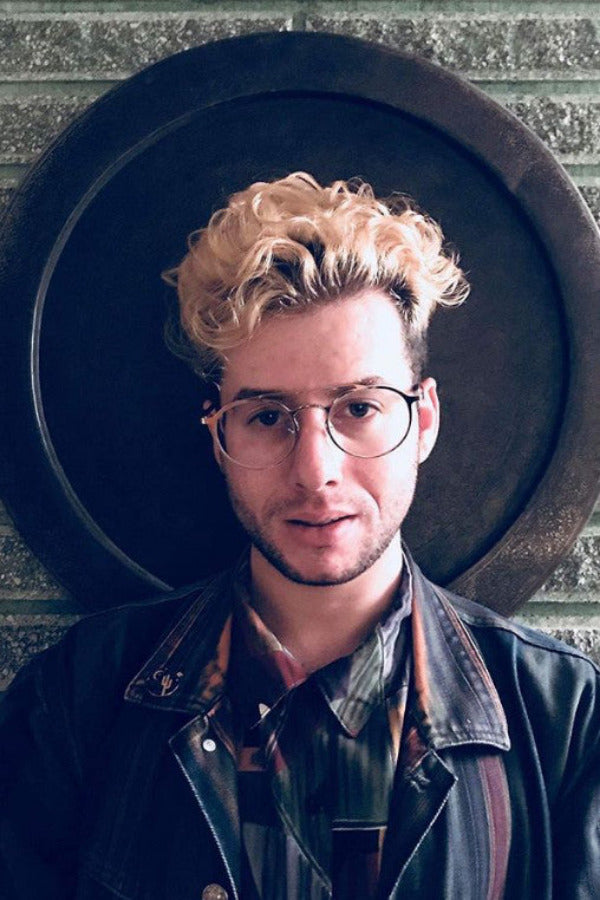Man with blonde hair and glasses wearing a dark jacket and plaid shirt against a circular black background.