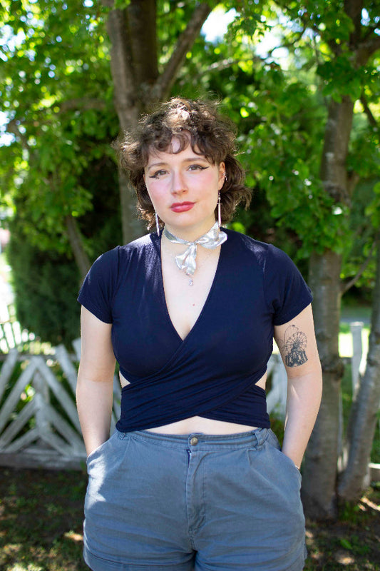 Woman wearing a navy blue top and light blue jeans standing outdoors with greenery in the background