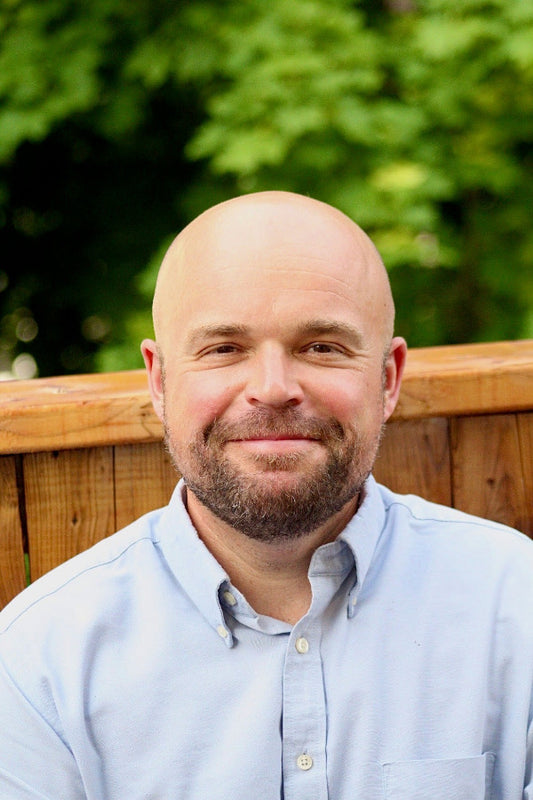 Man with a bald head and beard wearing a light blue shirt outdoors.