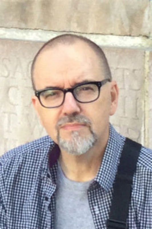 Man with glasses and a checkered shirt standing against a stone wall.