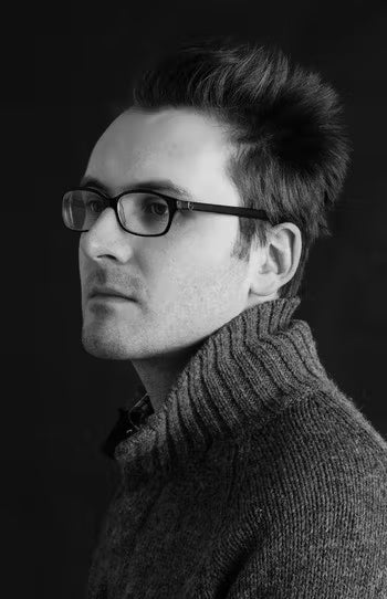 Black and white portrait of a man wearing glasses and a sweater against a dark background