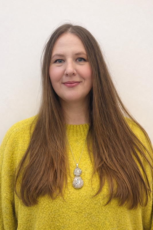 Lesley Goodyear, with long brown hair, a yellow sweater, and a medallion on a necklace. 