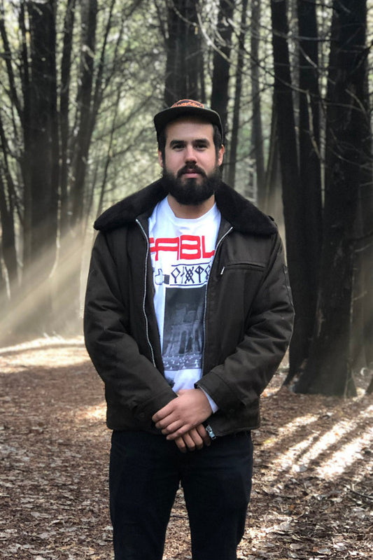 Man standing in a forest wearing a black jacket and white graphic t-shirt.