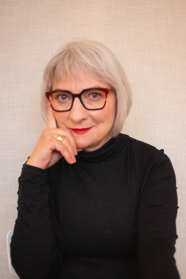Molly Peacock – a woman with gray hair and glasses wearing a black turtleneck against a beige background.