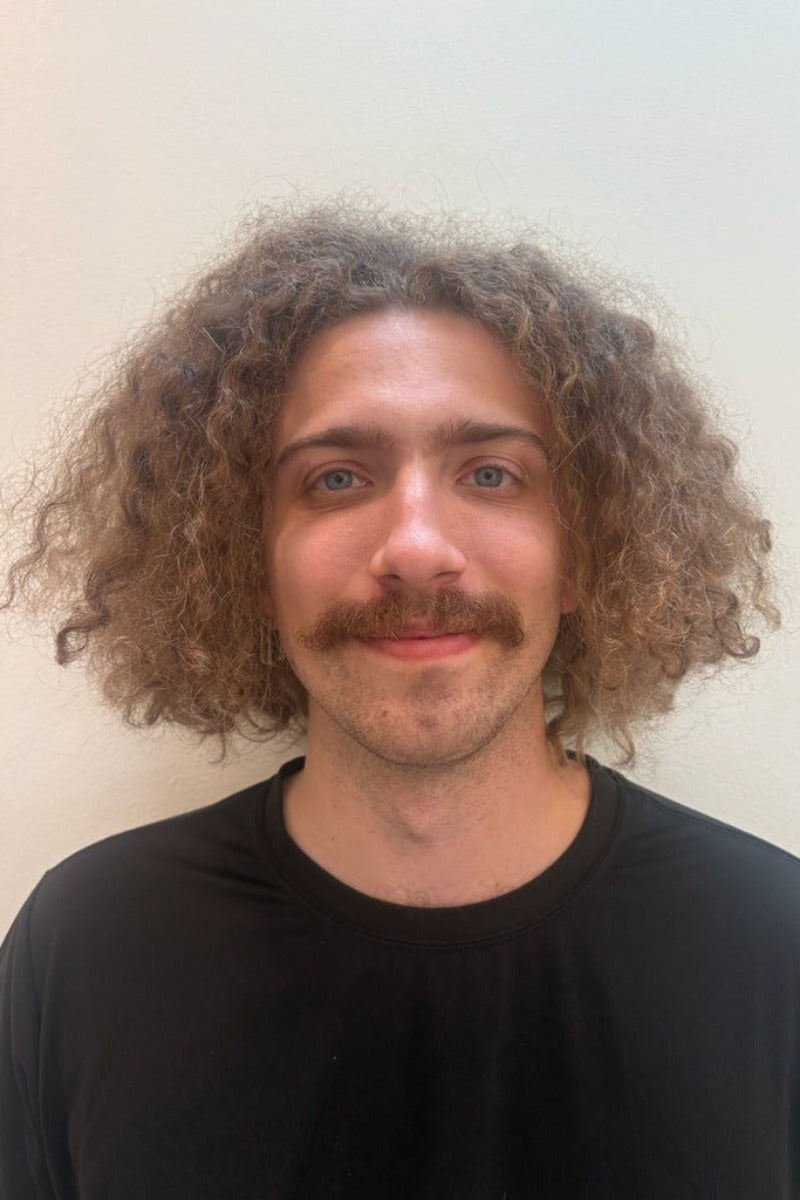 Sam Sturgeon, a man with curly brown hair and a mustache wearing a black t-shirt, photographed against a plain off-white wall.